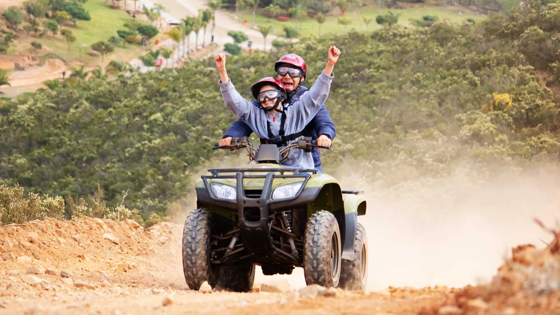 Two helmeted riders traveling on an ATV along a dusty Baja trail